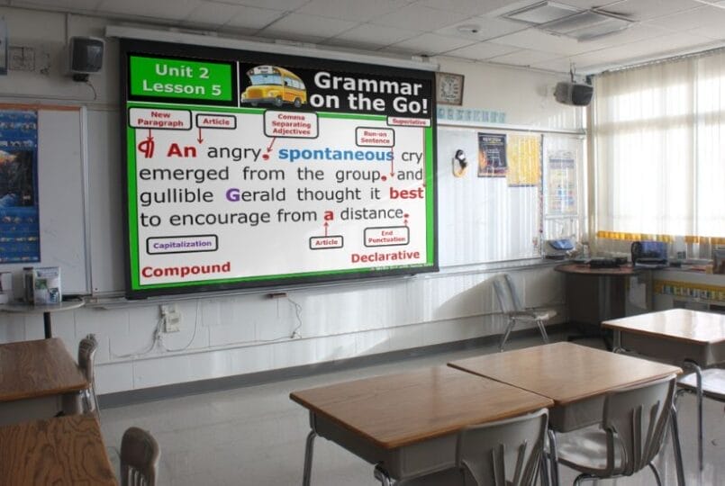 A classroom with empty desks and chairs faces a whiteboard displaying a colorful grammar lesson on sentence structure, featuring highlighted words and labels as part of the 9th grade English curriculum about spontaneous cry.
