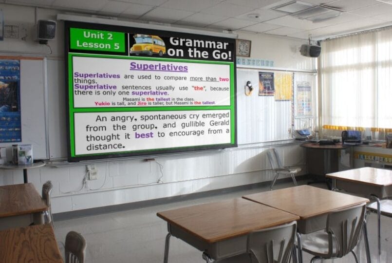 A classroom with empty desks faces a large screen displaying a superlatives lesson for Grammar on the Go! Unit 2, Lesson 5—perfect for grammar bell ringers middle school or engaging grammar warm-up activities. Sunlight streams in through the windows.