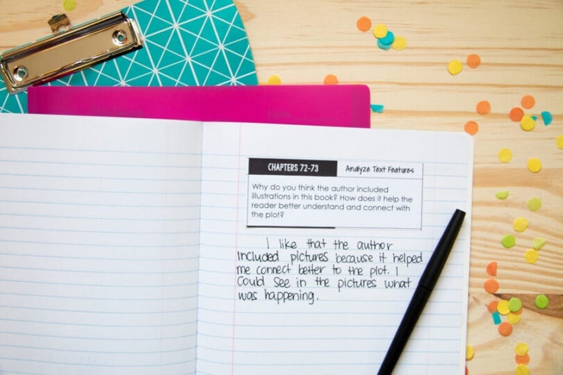 An open notebook displays a comprehension question about analyzing text features and a handwritten response, with a black pencil on the page. Nearby are a clipboard, pink folder, and colorful confetti on a yellow surface.