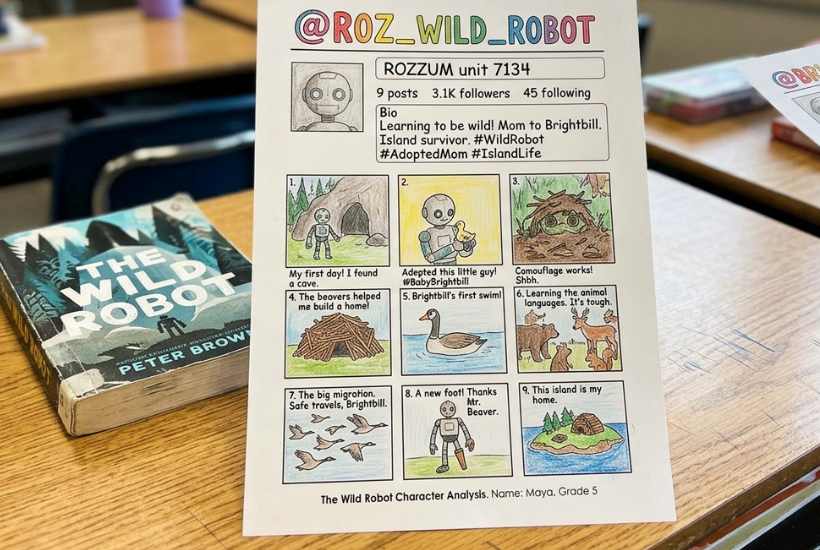 A worksheet titled @ROZ_WILD_ROBOT featuring a character summary of The Wild Robot, with colorful comic-style drawings and captions, lies on a desk beside the book—perfect for a Wild Robot novel study or classroom guide.